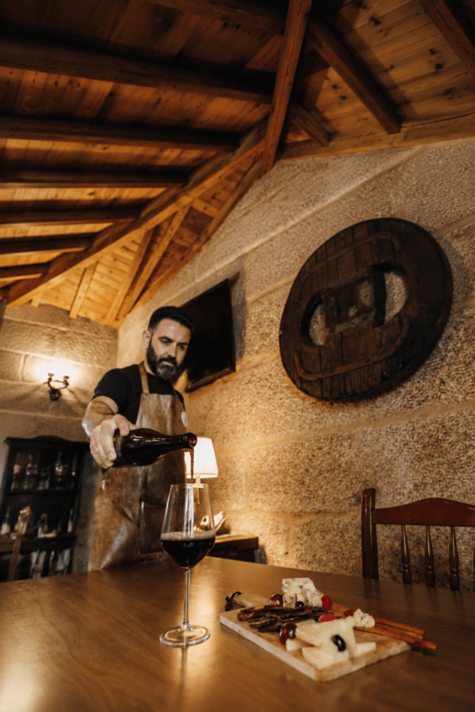 Propietario de Casa Nagarola sirviendo vino tinto en el comedor del restaurante, junto a una tabla de embutidos y quesos, en un ambiente rústico y acogedor en Fornelos de Montes, Galicia.