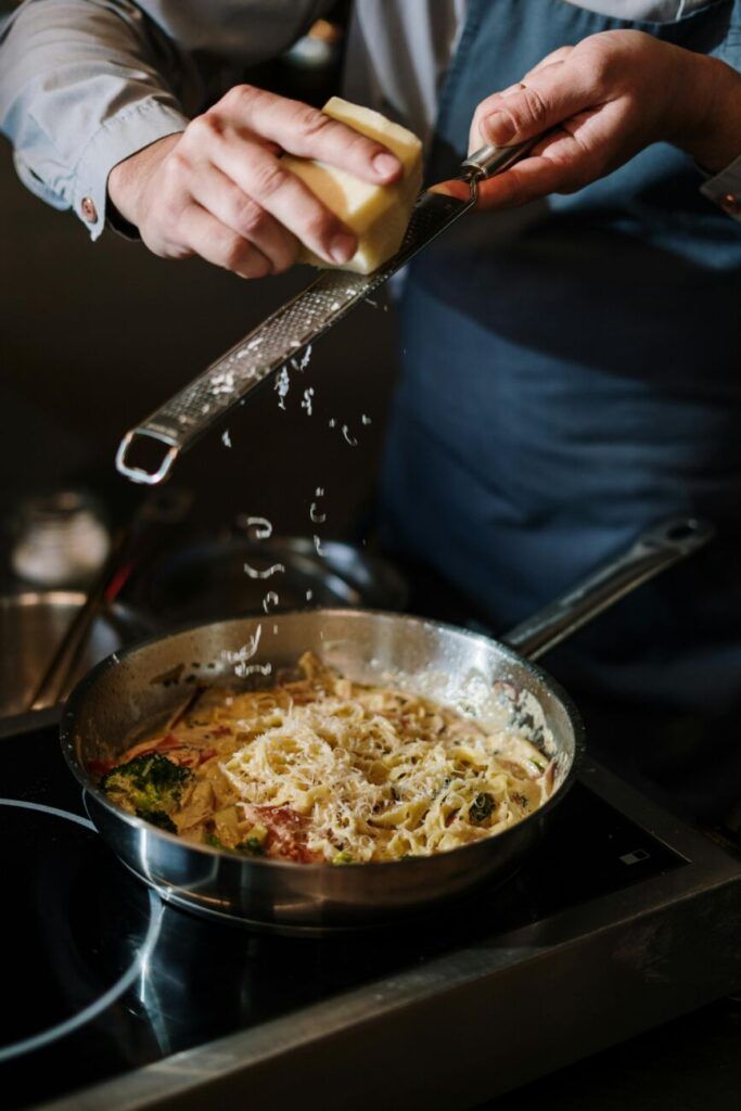 A chef in apron grates cheese onto pasta in a modern kitchen, capturing culinary artistry in action.