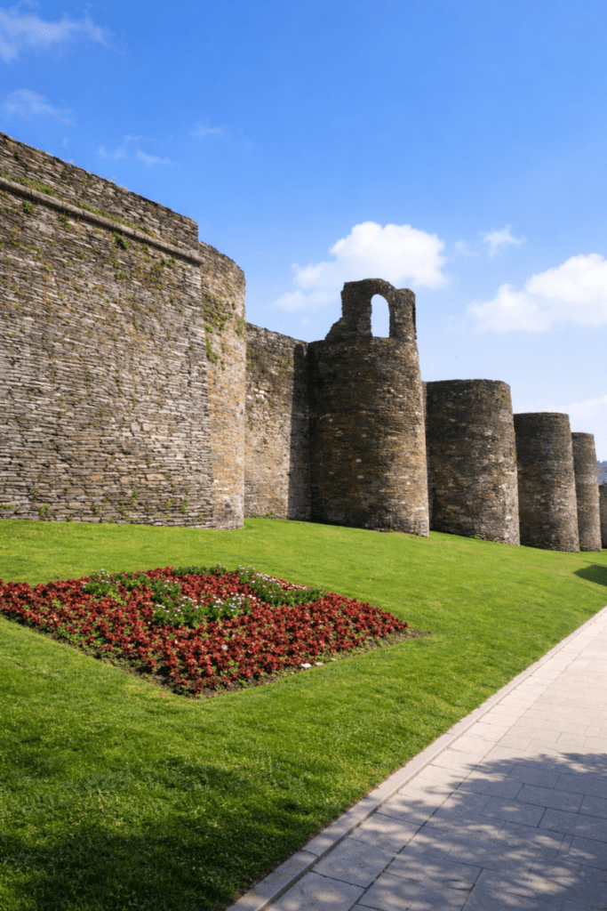 Muralla romana de Lugo en Galicia, con torres de piedra, césped y parterre de flores en primer plano, vista desde el paseo exterior en un día soleado.