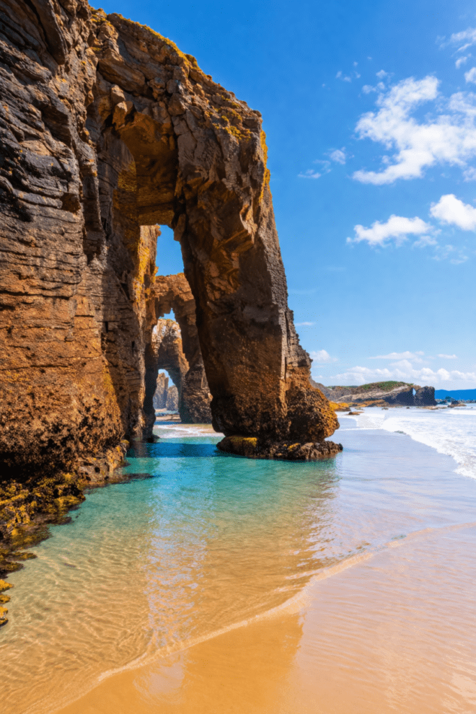 Playa de As Catedrais en Lugo, con impresionantes arcos de roca esculpidos por el mar, aguas turquesas y arena dorada en un día soleado.