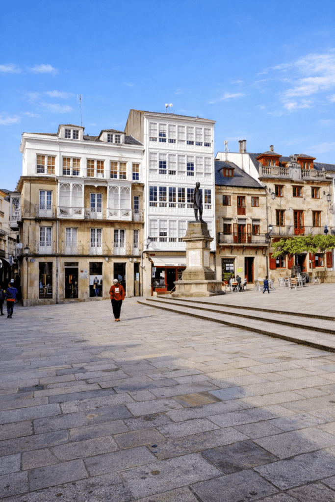 Plaza de Viveiro (Lugo) con edificios históricos de galerías blancas, estatua central y pavimento de piedra en un día soleado.