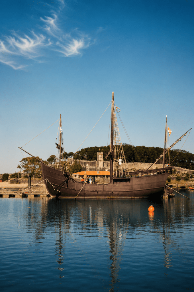 La carabela La Pinta en el puerto de Baiona al atardecer, con reflejos sobre el agua y la fortaleza de Monterreal al fondo.