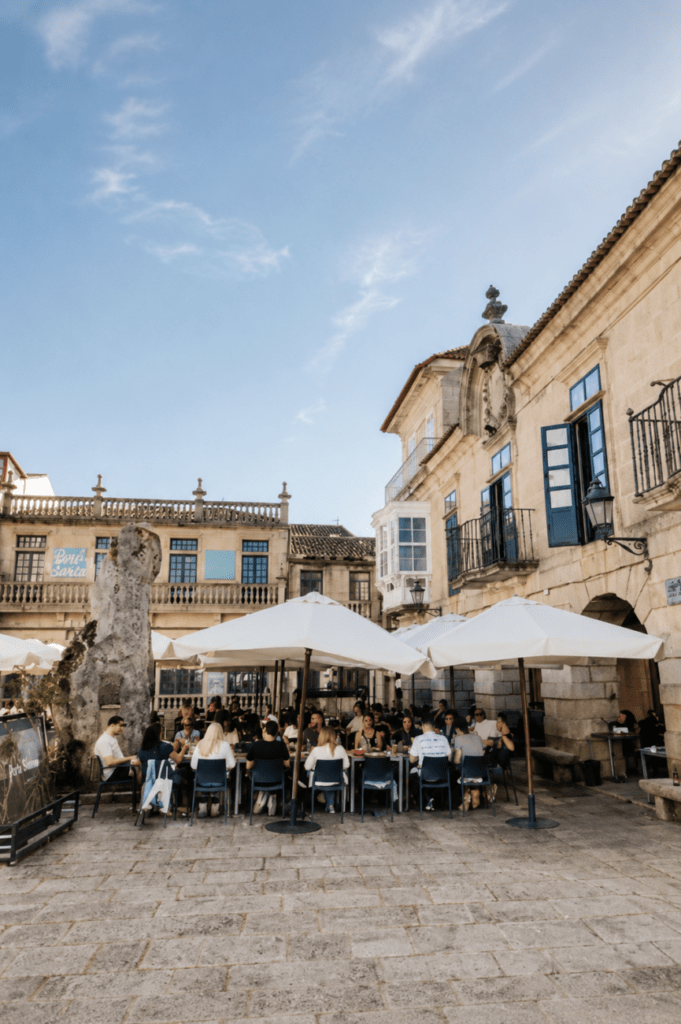 Terraza en plaza histórica de Baiona con edificios de piedra gallega, mesas bajo sombrillas blancas y ambiente elegante en un día soleado.