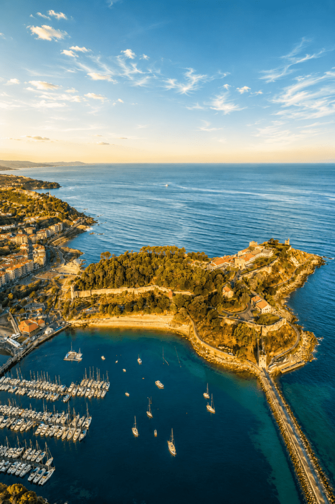 Vista aérea de Baiona al atardecer con la fortaleza de Monterreal, el puerto deportivo y el océano Atlántico iluminados por una cálida luz dorada.